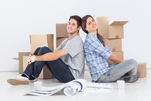 A young man and woman sit back-to-back on the floor of a bright, minimalist room surrounded by various cardboard boxes of different sizes, some sealed and others open. The man, with short dark hair, wears a light grey t-shirt, dark jeans, and casual sneakers, smiling comfortably as he leans against a stack of boxes. The woman, with long brunette hair, is dressed in a blue checkered shirt and grey jeans, looking upwards with a gentle smile. In front of them are packing materials, including rolled-up plans or posters, partially unrolled, along with two white mugs placed on the floor nearby. The background features a plain white wall, suggesting an indoor space preparing for a move or unpacking process typical of house removals in the Bayswater or central London area. The scene's natural lighting emphasizes the neutral tones of the boxes and furniture, indicating an organized move, consistent with professional removals services in London, such as those offered by Bayswater Man with Van.
