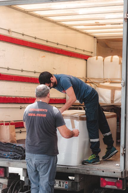 Two movers from Bayswater Man with Van are inside the back of a moving truck, with one standing on the truck bed and the other on the ground. They are handling large white packing blankets wrapped aro